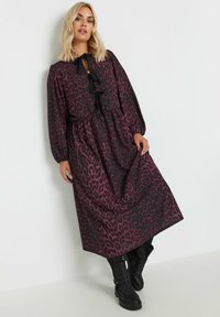 Pitkähihainen musta mekko, jossa on violetti leopardikuvio, vyötärösidonta ja napit, yhdistetty mustiin polvimittaisiin saappaisiin.