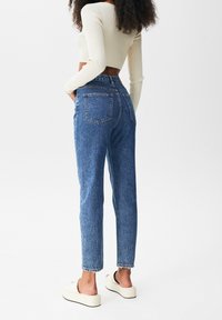 High-waisted blue denim jeans with a straight leg, frayed hem, and two back pockets, paired with a cream long-sleeve cropped top and white platform slides.