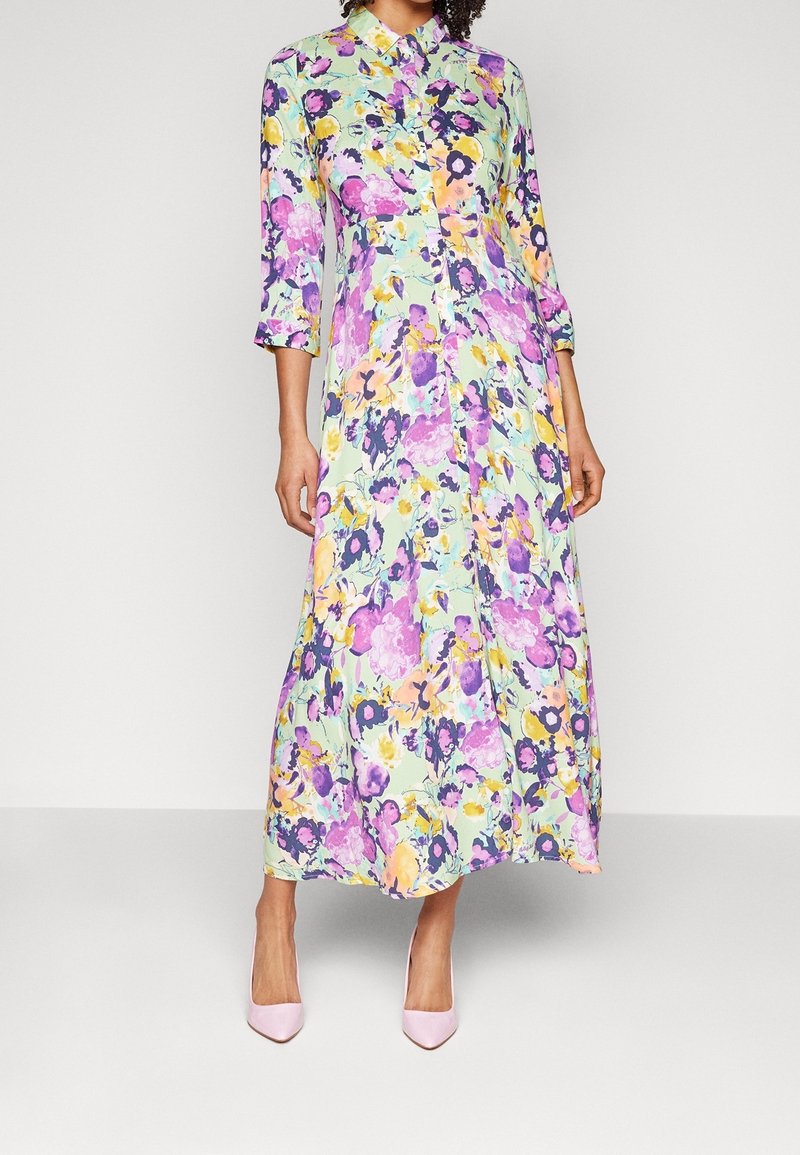 Pitkä kukkamekko vaaleanvihreänä, jossa on violetteja, keltaisia ja vaaleanpunaisia kuvioita. Mekossa on kaulus ja napitettu etuosa, sekä kolmen neljänneksen hihansuut.