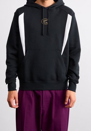 Person trägt schwarzen Hoodie mit weißen Seitenstreifen und einer Fronttasche, kombiniert mit locker sitzenden lila Hosen, vor einfachem Hintergrund.
