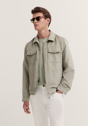 Light gray suede jacket with two chest pockets and a zipper, paired with a mint green shirt and white pants. Model wears sunglasses.