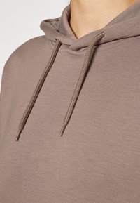 Sudadera con capucha marrón con tejido texturizado, bolsillo delantero y cordones trenzados con puntas metálicas. La capucha está bien definida.