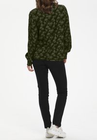 Chemise à manches longues vert olive avec un motif géométrique, présentant une coupe décontractée et des manches retroussées, associée à un pantalon noir ajusté.