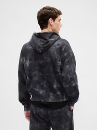 Homem com um hoodie em tie-dye preto e cinza e calças combinando, de perfil e a olhar para a direita contra um fundo simples.