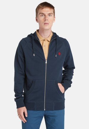 Uomo che indossa una felpa blu navy con zip sopra una polo beige e pantaloni blu, con una mano nella tasca della felpa, in piedi contro uno sfondo neutro.