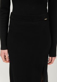 Robe en tricot noire à manches longues, avec une taille plissée et une fente sur le côté. Agrémentée d'un petit détail en métal doré.