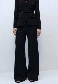 Blazer nero su misura con bottoni, abbinato a pantaloni neri a gamba larga. Presenta una cintura in vita, linee pulite e una texture liscia.