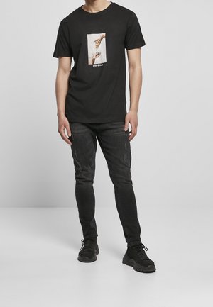 T-shirt noir avec un motif graphique de deux mains, l'une tendant vers l'autre, porté avec un jean foncé et des baskets noires.