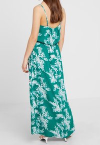 Grön maxiklänning med vitt blommönster, justerbara spaghetti-stövlar, elastisk midja och flödande tyg. Bakdelen har en liten öppning.