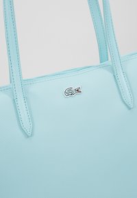 Sac fourre-tout bleu clair en matériau texturé, avec deux longues poignées, arborant un petit logo de crocodile brodé dans une couleur contrastante.