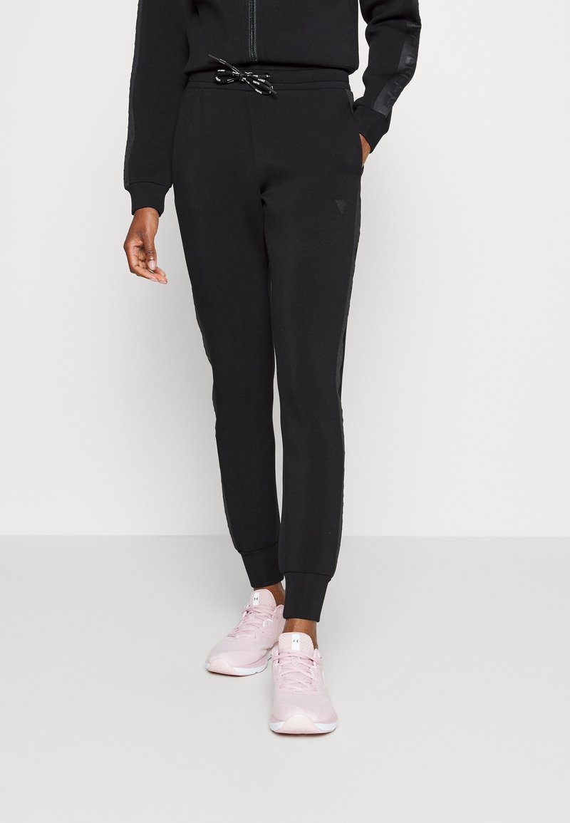 Svart joggebukser laget av mykt stoff, med elastisk midjebånd og svart snor, smalner inn ved anklene og har sidelommer.