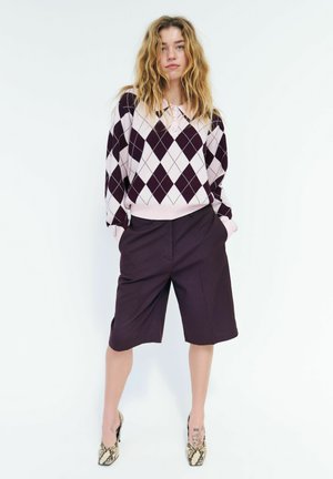 Junge Frau mit lockerem blondem Haar trägt einen pink-schwarzen Argyle-Pullover, schwarze knielange Shorts und High Heels aus Schlangenleder.