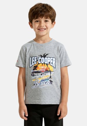 Jeune garçon souriant aux cheveux bruns portant un t-shirt gris avec un motif coloré de coucher de soleil à la plage et un palmier, un pantalon noir, debout sur un fond blanc.