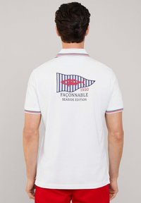Polo blanco con cuello a rayas en rojo y azul marino. La parte trasera presenta un diseño de bandera azul y blanca con un gráfico de pez rojo y texto.