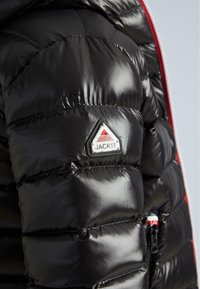 Jachetă puffer neagră cu finisaj glossy, având secțiuni quilted și o etichetă logo triunghiulară inscripționată "JACKIT". Accente de fermoar roșu vizibile.