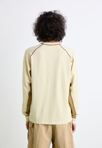 Long-sleeve beige sweater with a textured fabric and brown side accents. Features a crew neck and raglan sleeves.