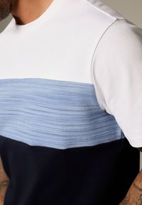 Kortärmad t-shirt med vit överdel, ljusblå texturerad rand i mitten och mörkblå nederdel. Slät tyg med lager-på-lager-design.