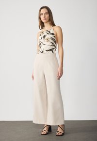 Blouse sans manches avec col, imprimé floral dans des nuances de crème et de noir, associée à un pantalon large beige clair et des talons aiguilles noirs à lanières.