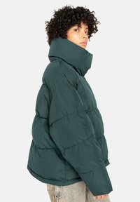 Persoon met krullend haar, gekleed in een donkergroene oversized pufferjack met een hoge kraag, gecombineerd met lichtgekleurde jeans.