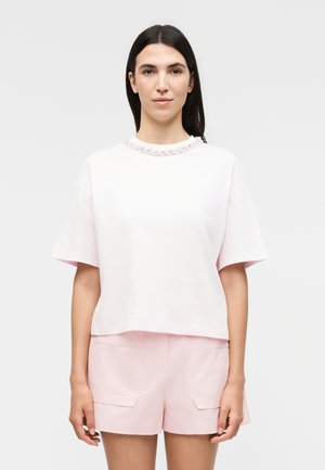 Γυναίκα με ίσια μαύρα μαλλιά, φορώντας λευκό κοντομάνικο τοπ με λουριά στο λαιμό και ανοιχτό ροζ σορτς σε απλό φόντο.