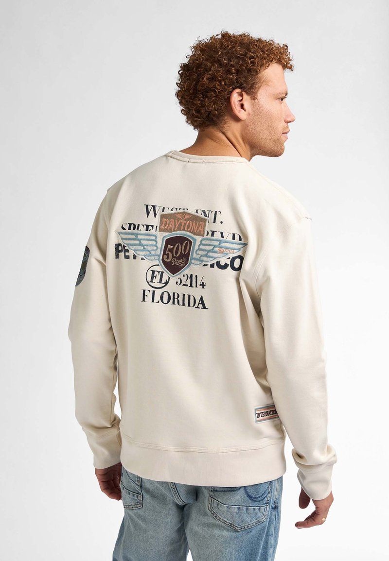 Felpa crema con stampe grafiche sulla schiena che presenta la dicitura "Daytona 500" e disegni di ali. Polsini e orlo a costine, abbinata a jeans blu.