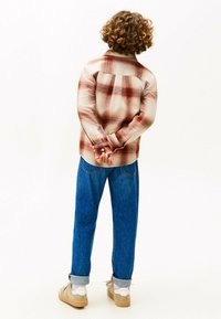 Camicia a quadri nei toni beige, rosso e arancione, con taglio dritto. Jeans in denim blu, leggermente arrotolati ai polsini. Sneakers beige con suole bianche.
