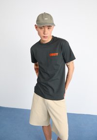 Dark gray t-shirt with red "OBEY" logo, paired with beige plaid shorts. The cap is light gray with white "OBEY" text.