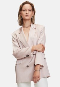 Dobbeltbrystet blazer i lys pink satin med slag og sorte knapper, der har en afslappet pasform og sidelommer. Båret over hvide bukser.