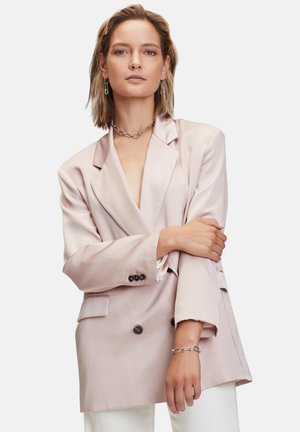 Blazer croisé en satin rose clair avec revers et boutons noirs, offrant une coupe décontractée et des poches latérales. Porté avec un pantalon blanc.