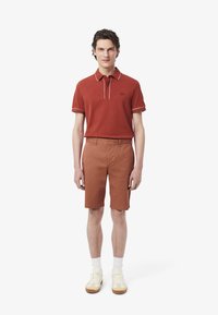 Polo rosso con bordi bianchi, abbinato a pantaloni corti color ruggine. Il modello indossa calze bianche e sneakers beige. Design semplice, vestibilità casual.