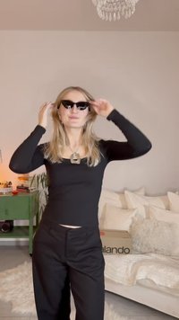 Occhiali da sole neri con design angolare, indossati con una maglia nera a maniche lunghe e pantaloni neri. Collana argentata spessa e pendente notable.