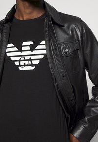 Svart läderjacka över en svart t-shirt med en vit örnlogotyp och horisontella ränder. Jackan har en ficka med knappar.