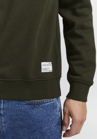 Højre side af en person iført en olivengrøn sweatshirt med et hvidt mærke nær kanten og blå denim jeans, hånd afslappet ved deres side.