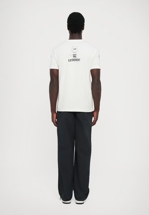 T-shirt en coton blanc avec le texte "EXTROVERT" et un graphique au dos, associé à un pantalon noir à jambes larges et des baskets blanches.