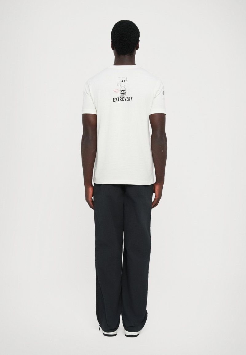 Camiseta de algodón blanca con el texto y gráfico "EXTROVERT" en la parte posterior, combinada con pantalones de pierna ancha negros y zapatillas blancas.
