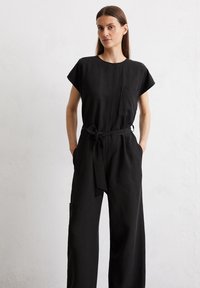 Zwarte jumpsuit met korte mouwen, een zak en een tailleband. Gemaakt van een lichtgewicht stof met een relaxte pasvorm en wijde benen.