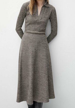 Femme portant une robe en maille marron texturée à manches longues, col, taille cintrée et jupe évasée, debout devant un fond clair.