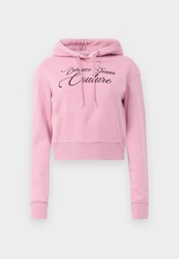 Hoodie corto rosa con cappuccio regolabile e la scritta "Versace Jeans Couture" in glitter nero sulla parte frontale. Tessuto morbido, orlo a costine.