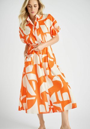 Vrouw die de riem aanpast van een halflange jurk met korte mouwen en een patroon in oranje en wit, met een kraag, staand tegen een effen achtergrond.