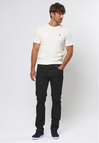 Witte shirt met korte mouwen en ribbeltextuur, zwarte slim-fit jeans en blauwe sneakers. De look is casual met minimale details.