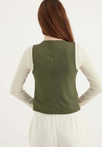 Olivgrön väst i slätt tyg, figursydd design med rund halsringning och utan knäppningar, bärs över en långärmad beige topp.
