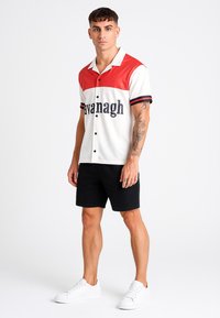 Camisa de botones de manga corta con paneles rojos y blancos, con acentos negros y la palabra "avanagh" en fuente negrita. Combinada con pantalones cortos negros.