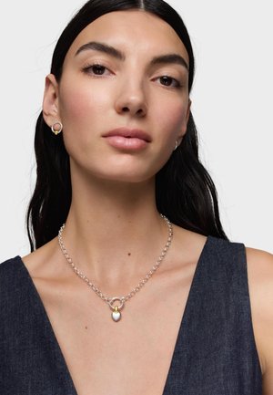 Kobieta z długimi ciemnymi włosami nosząca srebrny łańcuszek z małą zawieszką w kształcie serca w kolorach złota i srebra oraz pasujące kolczyki koła, ciemny, bezrękawnik.