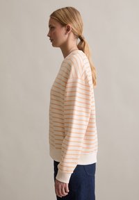 Pomarańczowo-biała bluza w paski o luźnym kroju, z prążkowanymi mankietami i miękką teksturą, zestawiona z ciemnoniebieskimi dżinsami.