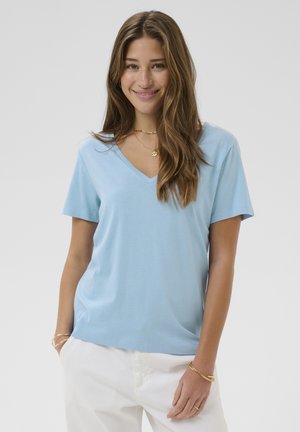 T-shirt bleu clair à manches courtes et col en V, en coton doux, avec une coupe décontractée et une texture lisse. Porté avec un pantalon blanc.