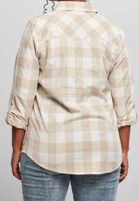 Persona che indossa una camicia a maniche lunghe beige e bianca a scacchi con le maniche arrotolate e jeans blu, vista da dietro.