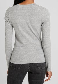 Eine Person, die ein eng anliegendes, hellgraues Langarmshirt und schwarze Jeans trägt, von hinten vor einem schlichten grauen Hintergrund gezeigt.