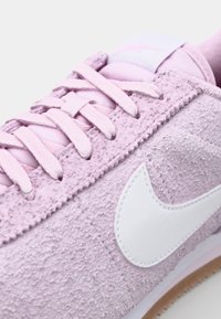 Pink textured sneaker with fabric upper, white Nike swoosh, and matching laces. Light-colored sole with a subtle grip pattern.