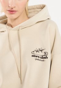 Beige hettegenser med en frontlomme, prydet med et svart grafisk design av stjerner og en sikkerhetsnå. Laget av bomull med snøring.