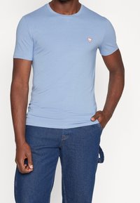 Lys blå bomulds t-shirt med korte ærmer, rund halsudskæring og lille rødt trekantet logo på brystet; parret med mørkeblå denim jeans.